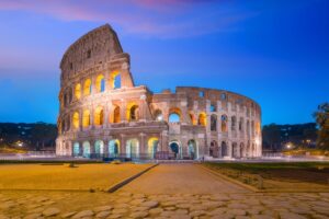 The image captures the Colosseum in Rome at dusk, a prime holiday destination in Europe. A clear sky transitions from blue to warm pink, illuminating the ancient amphitheater's arches and ruins. The cobblestone pavement in the foreground hints at timeless travel tips for history lovers.